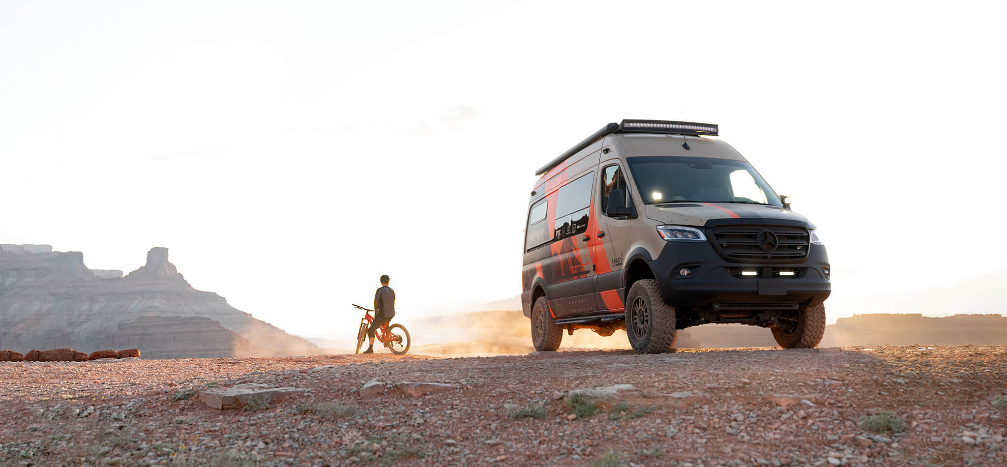A mountain biker looks out over the desert beside a custom Sprinter camper van.