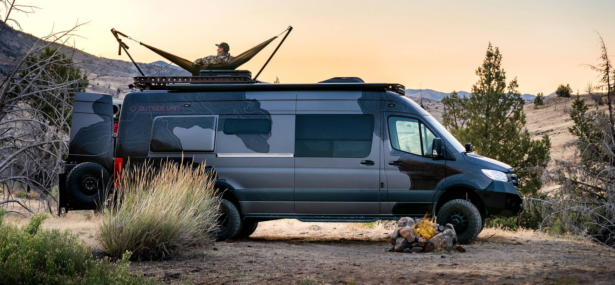 A hunter rests on a hammock on top of his 4x4 Sprinter van, looking out over the surrounding grasslands. A campfire burns in a firepit next to the van.