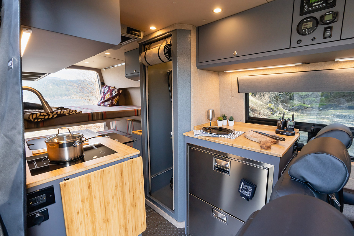 Van interior with fully enclosed aluminum shower, stainless steel refrigerator, and grey overhead cabinetry