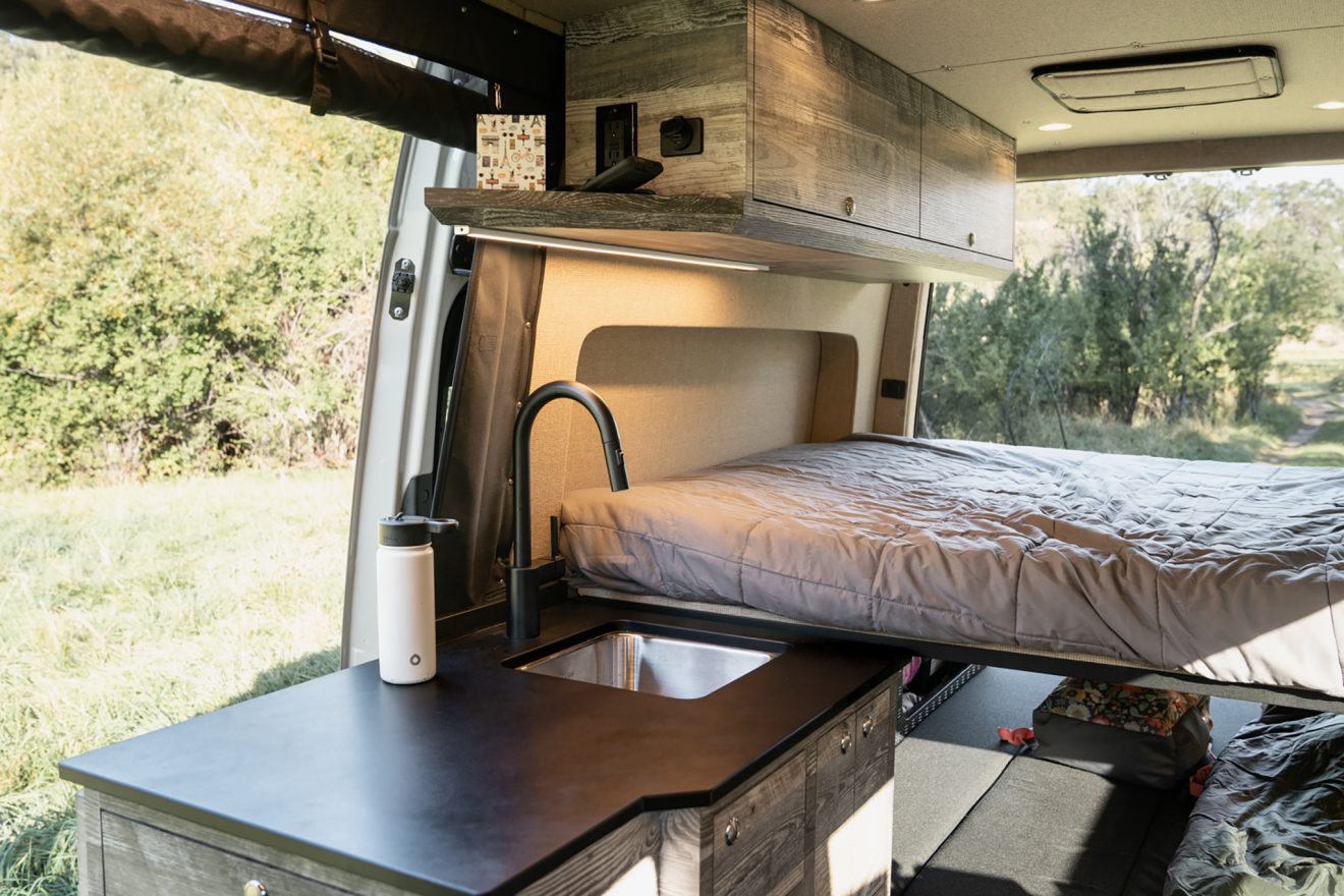 Van interior with galley kitchen, raised bed panel, and overhead cabinetry