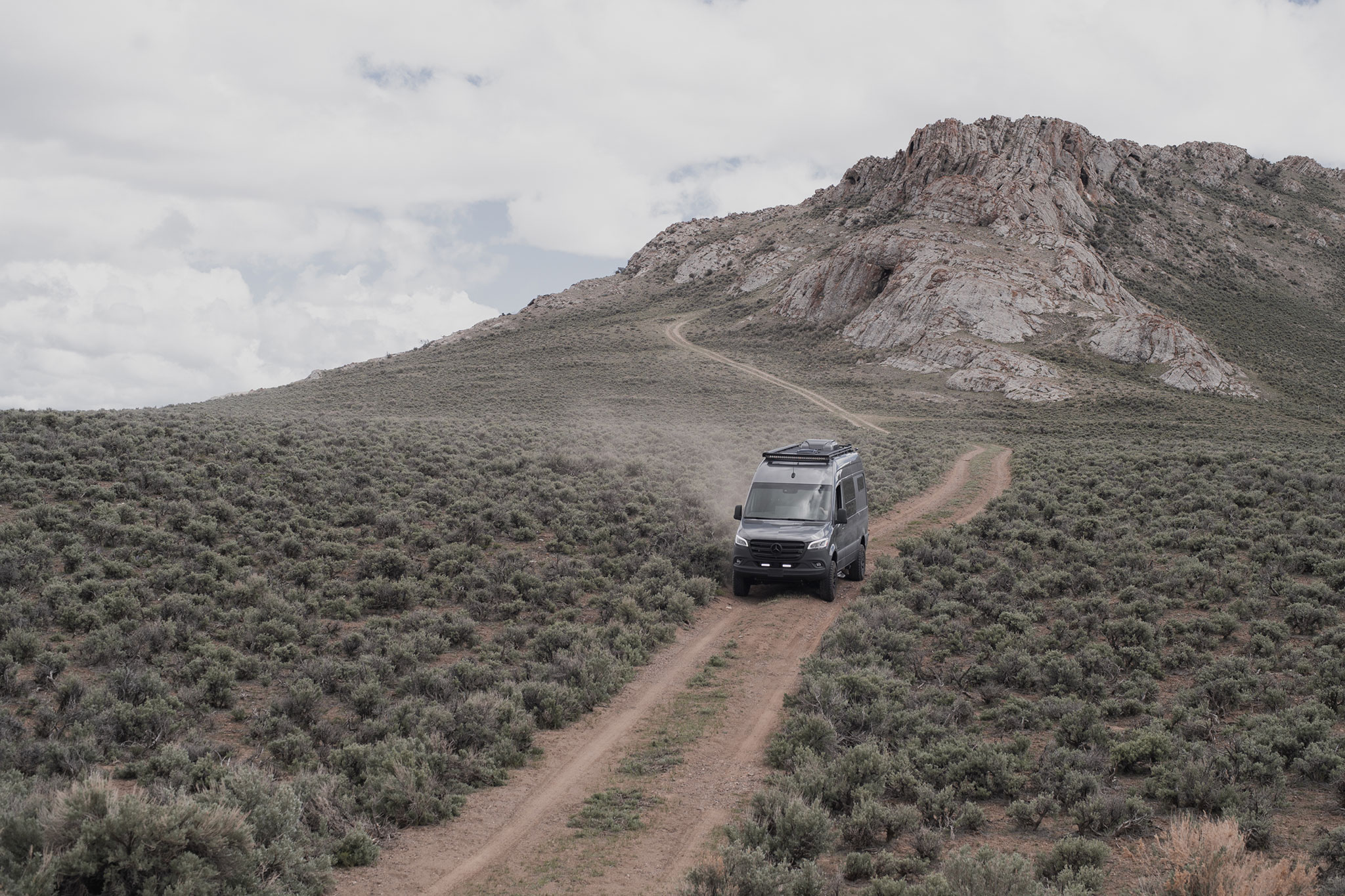A customized Sprinter van drives down a dirt trail with a rocky outcropping in the distance behind it.